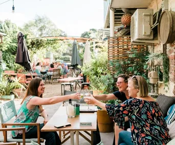 Three women toast with wine glasses at an outdoor restaurant, surrounded by green plants and relaxed atmosphere.