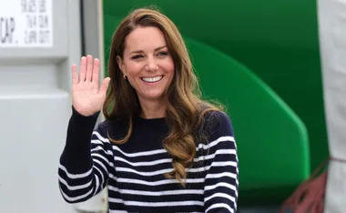 "Person in navy striped sweater waves and smiles warmly against a green and white background."