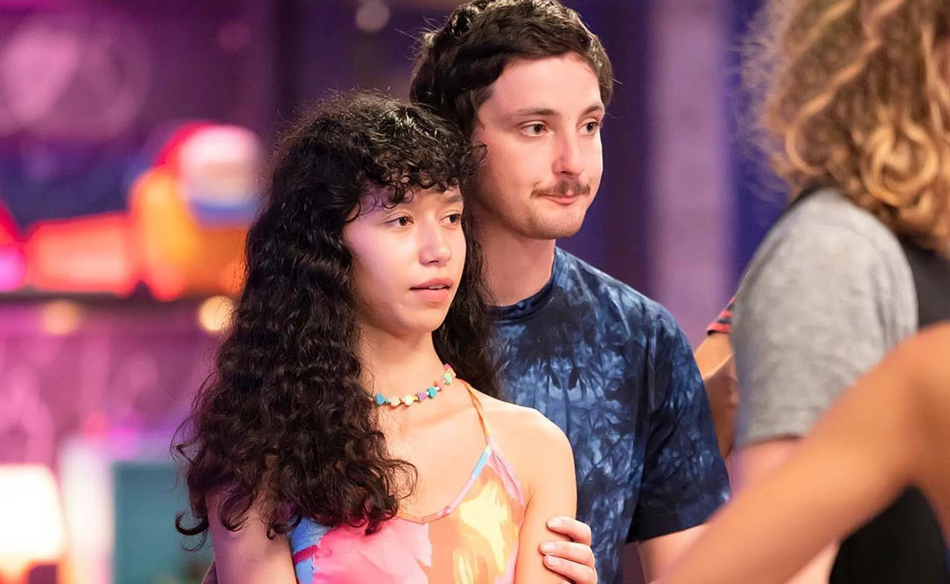 Two people stand close together, one with long curly hair and a colorful dress, the other with short hair and a blue shirt.
