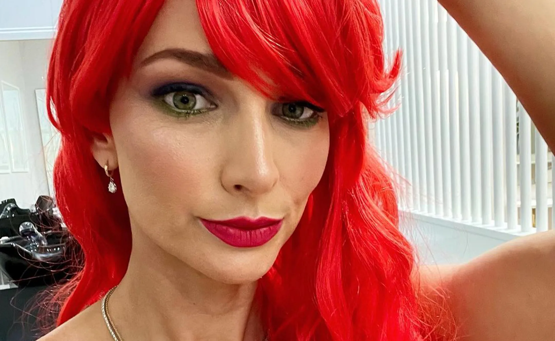 Red-haired woman with bold makeup, wearing a pendant necklace and drop earrings, in a brightly lit room.