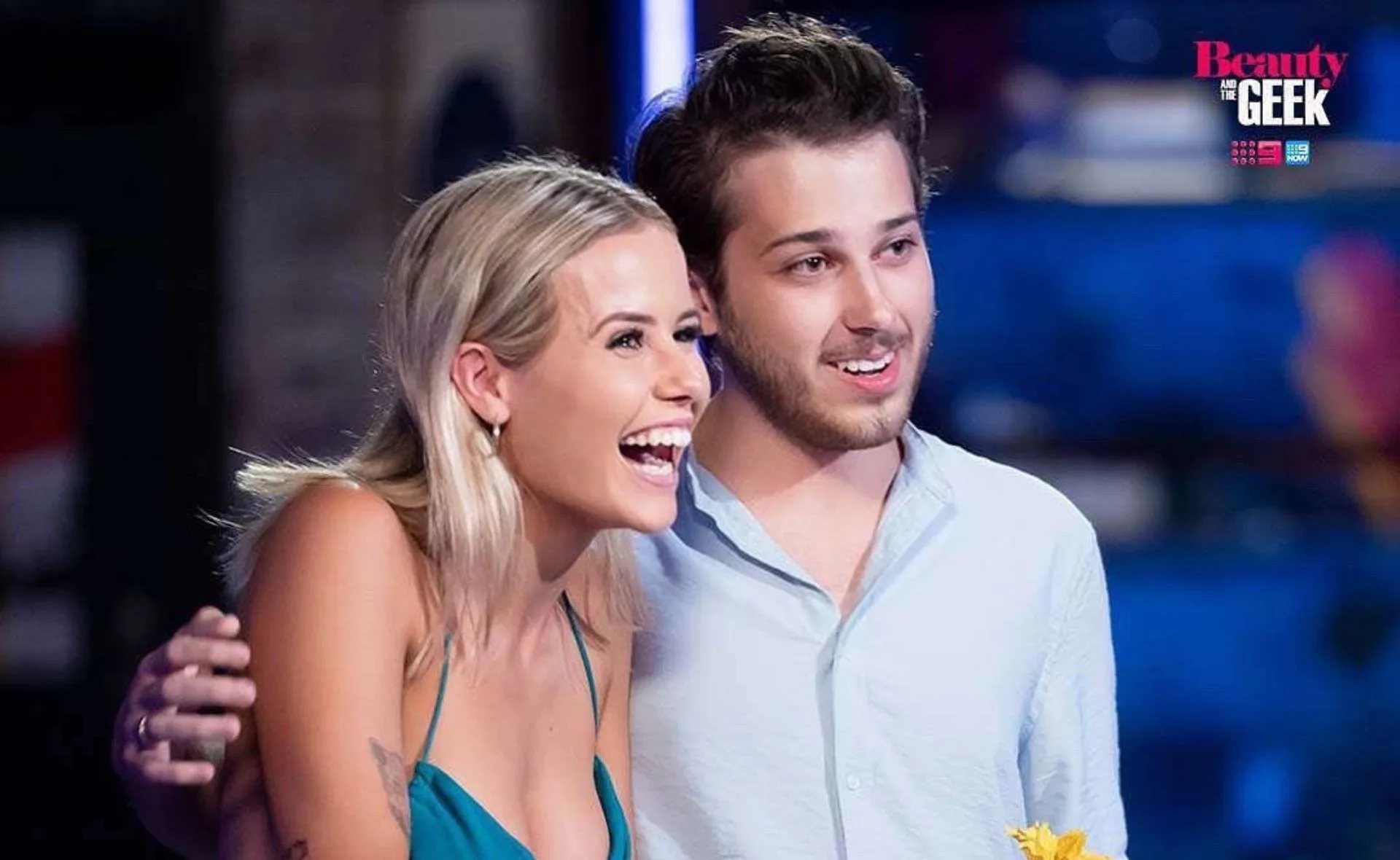 Contestants on "Beauty and the Geek" smiling and embracing, wearing casual outfits. Logo in background.