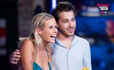 Contestants on "Beauty and the Geek" smiling and embracing, wearing casual outfits. Logo in background.