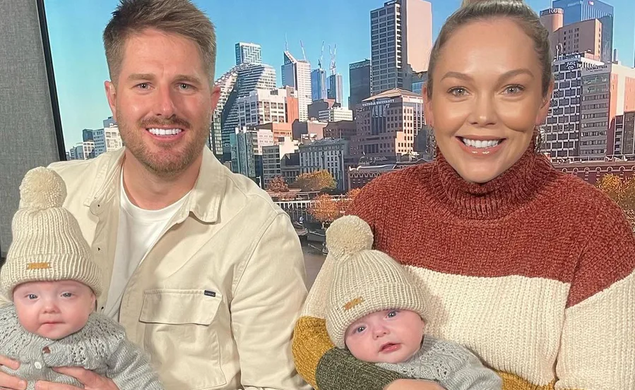 Family portrait with a couple holding twin infants in knit hats, cityscape in background.