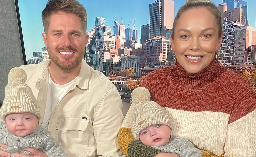 Family portrait with a couple holding twin infants in knit hats, cityscape in background.