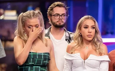 Three individuals appear on a TV set, one woman looks emotional, wiping her eye, while the others look serious.
