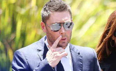 Man in suit and sunglasses touching his chin, with greenery in the background.