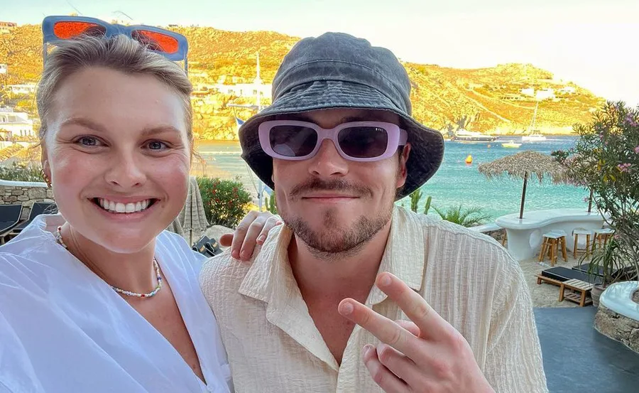 A smiling couple takes a selfie at a sunny seaside location, with boats and hills in the background.