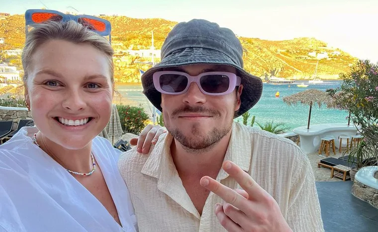 A smiling couple takes a selfie at a sunny seaside location, with boats and hills in the background.