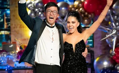 Two elated winners in formal attire celebrate at the "Beauty and the Geek Australia 2022" event, amidst colorful balloons.