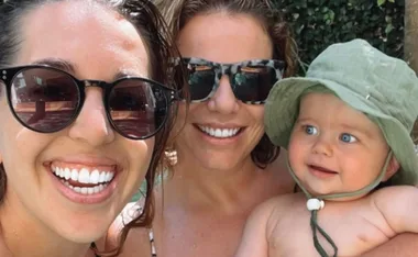 Two women in sunglasses smiling with a baby in a green hat, outdoors.
