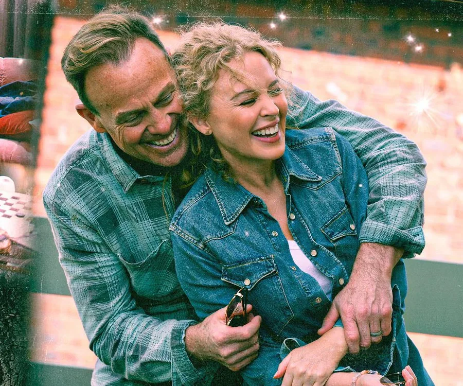 A smiling couple in denim embraces outdoors, background of brick and stars, conveying joy and affection.