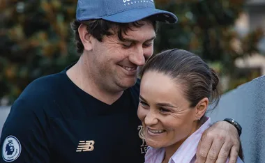 A couple smiling outdoors, with one person wearing a cap and the other with hair tied back.