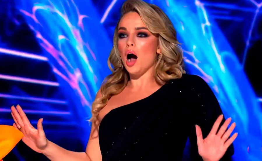 Woman in a black dress expresses shock; colorful background on "The Masked Singer."