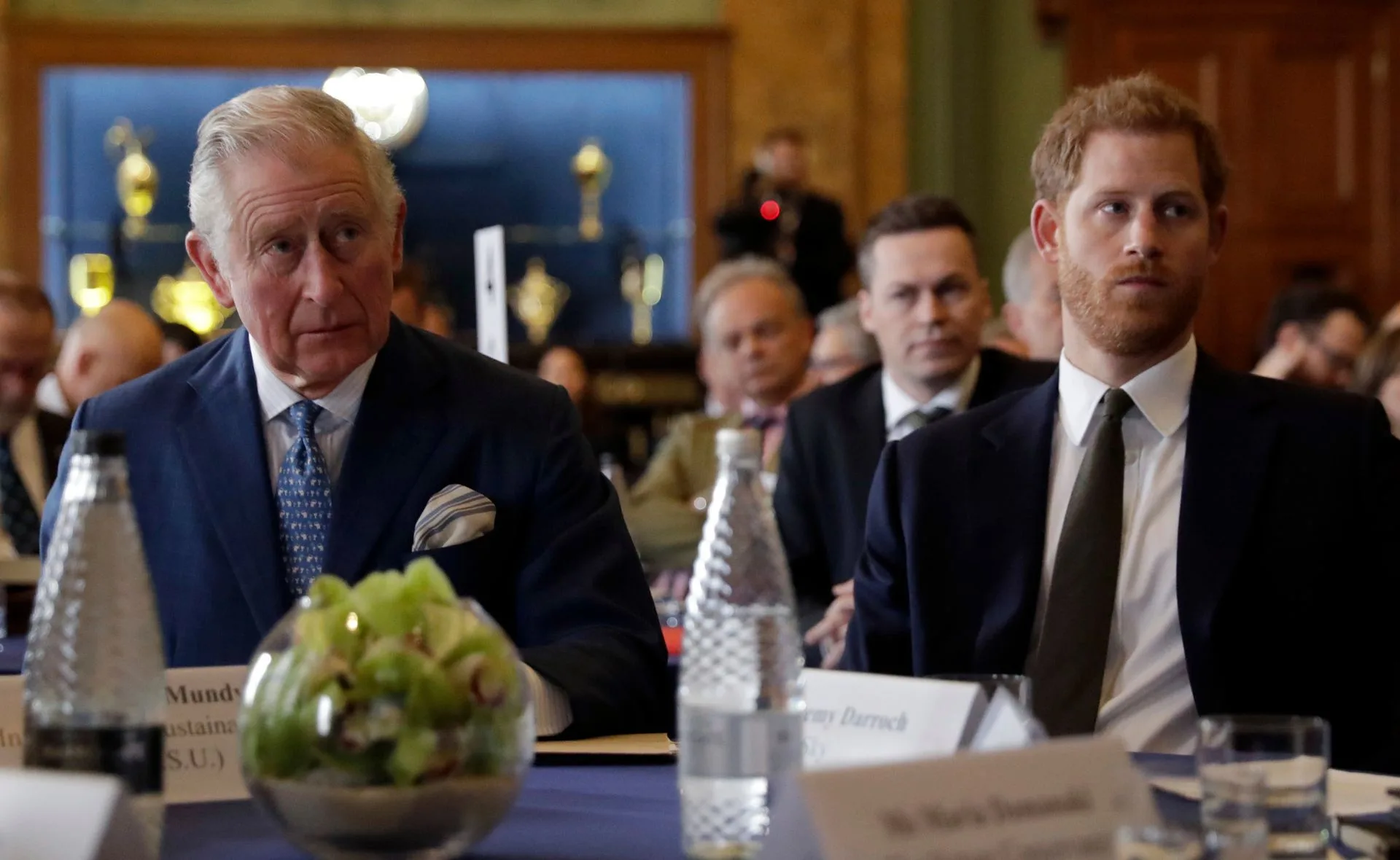 Two men in suits sitting at a conference table with others in the background.