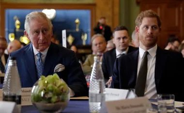 Two men in suits sitting at a conference table with others in the background.