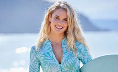Blonde woman smiling on the beach, holding a surfboard, wearing a light blue patterned swimsuit.