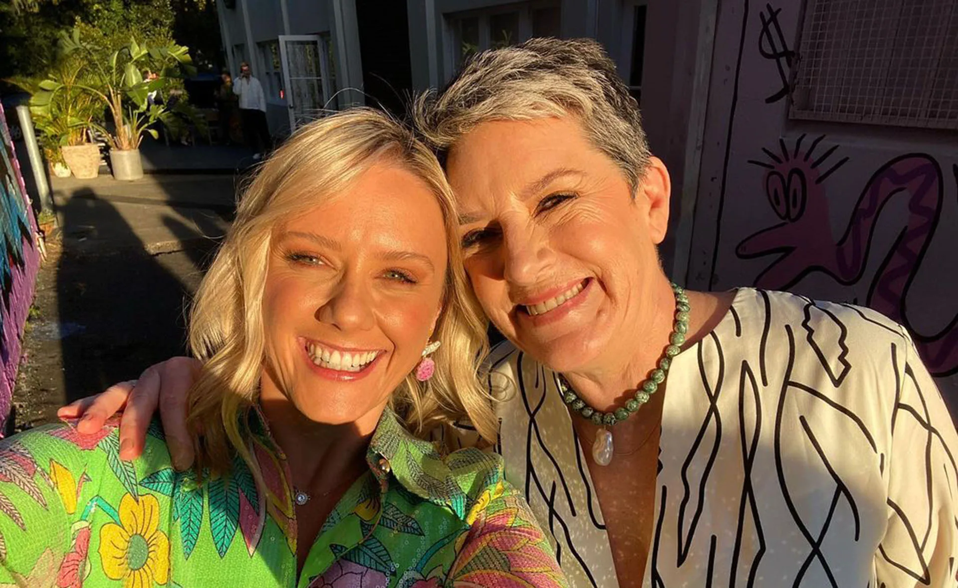 Two women smiling in a sunny outdoor setting, with colorful wall art in the background.