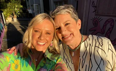 Two women smiling in a sunny outdoor setting, with colorful wall art in the background.
