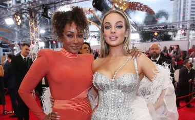 Two women posing on a red carpet, one in an orange dress and the other in a silver embellished gown.