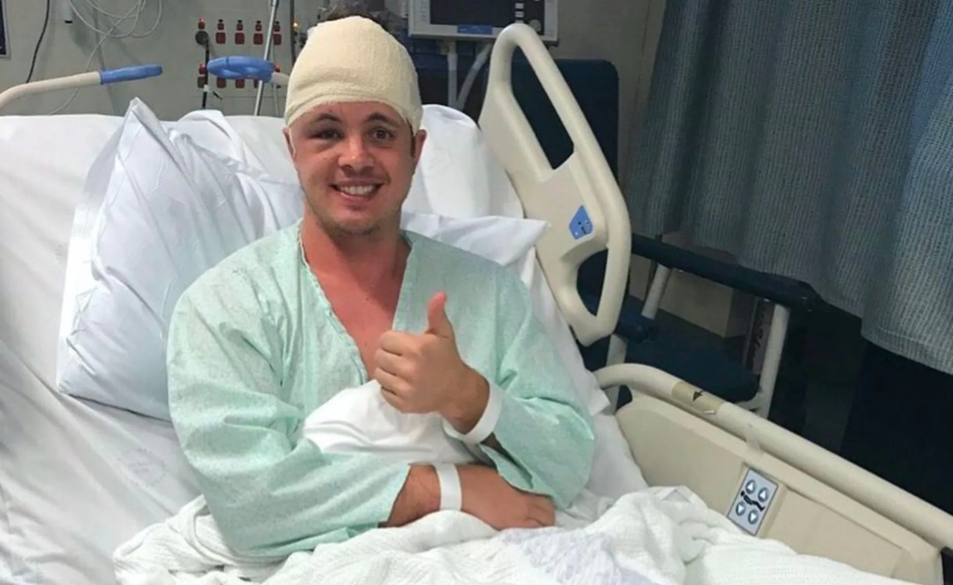 A man in a hospital bed with a head bandage, giving a thumbs up and smiling.