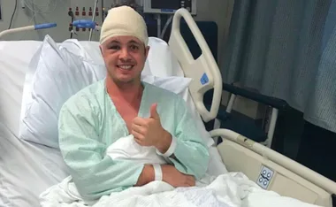 A man in a hospital bed with a head bandage, giving a thumbs up and smiling.