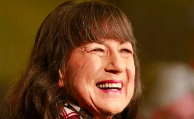 A woman with long brown hair and a bright smile, wearing a patterned outfit, against a blurred background.