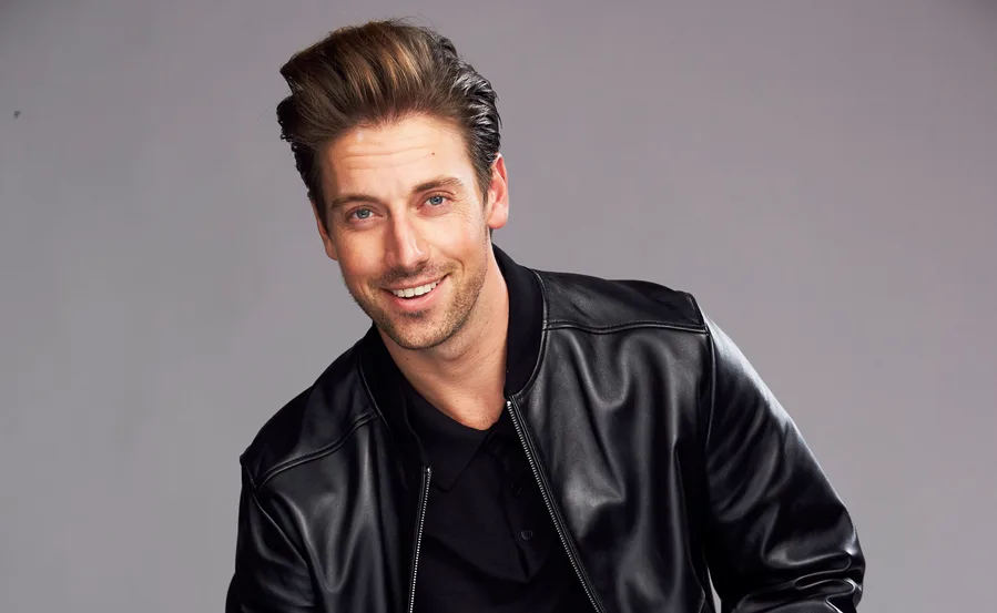 Man in a black leather jacket smiling, standing against a gray background.