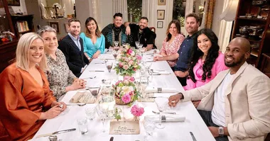 “Group of people sitting around a table with floral centerpiece, smiling and having dinner in a warmly lit room.”