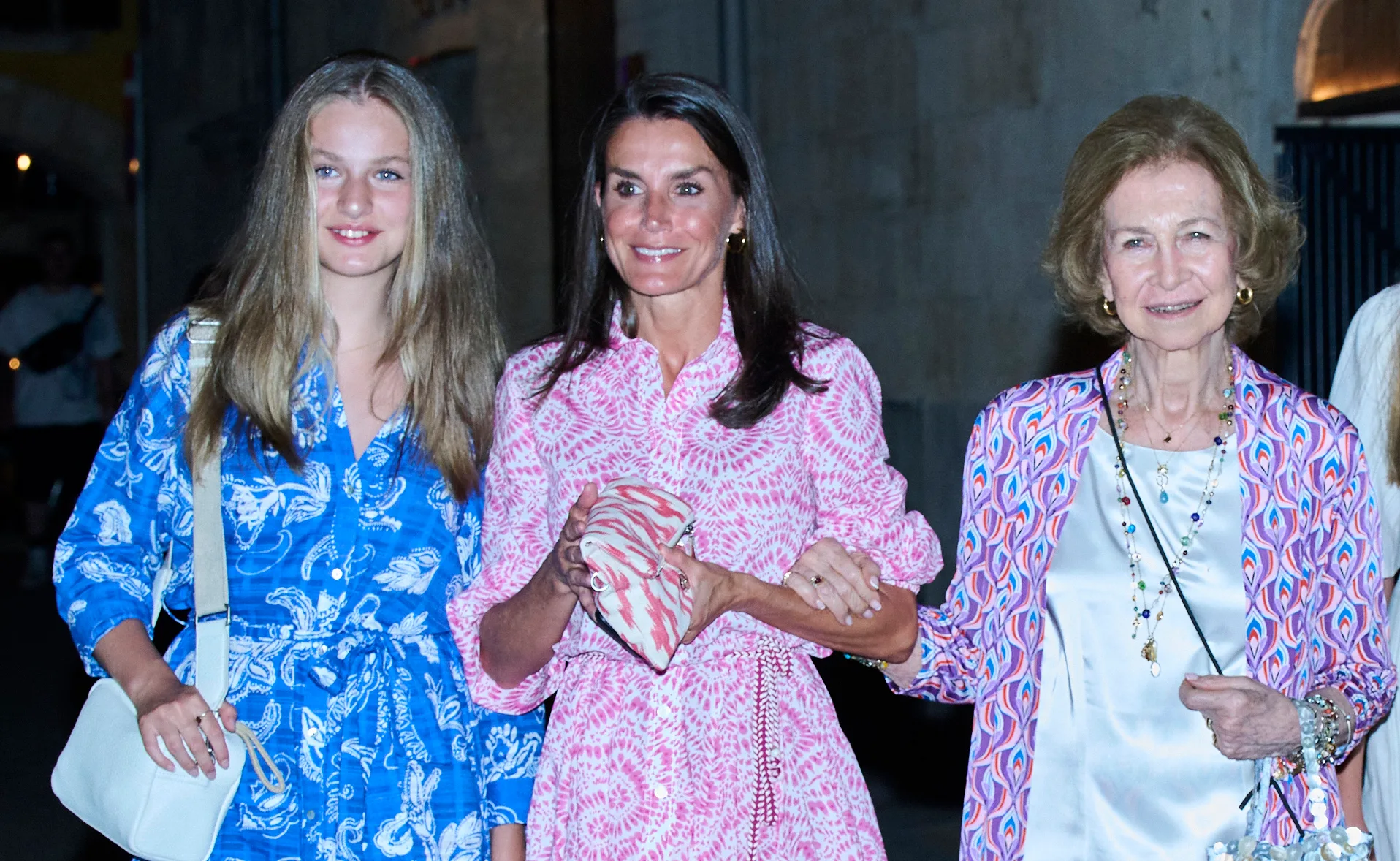 Three women in colorful dresses smiling at an evening event, one in pink and two in blue patterns.
