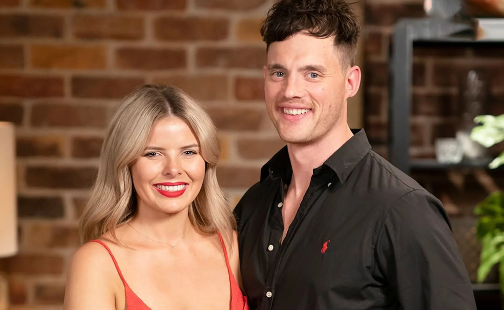 A smiling couple stands in front of a brick wall, both in casual attire.