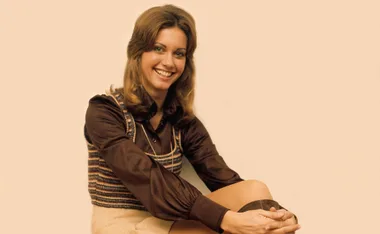 Olivia Newton-John smiling, seated, wearing a brown blouse and striped vest against a plain background.