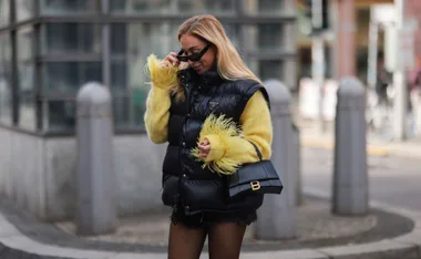 A woman in a black puffer vest and yellow sweater with feather cuffs, holding a black handbag, adjusts her sunglasses.