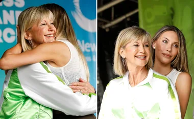 Two women embracing and smiling on stage at an event wearing casual and sporty clothing with a green and blue backdrop.
