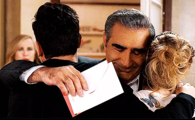 A man hugging a woman and another person, holding an envelope with a contented expression, with a person in the background.