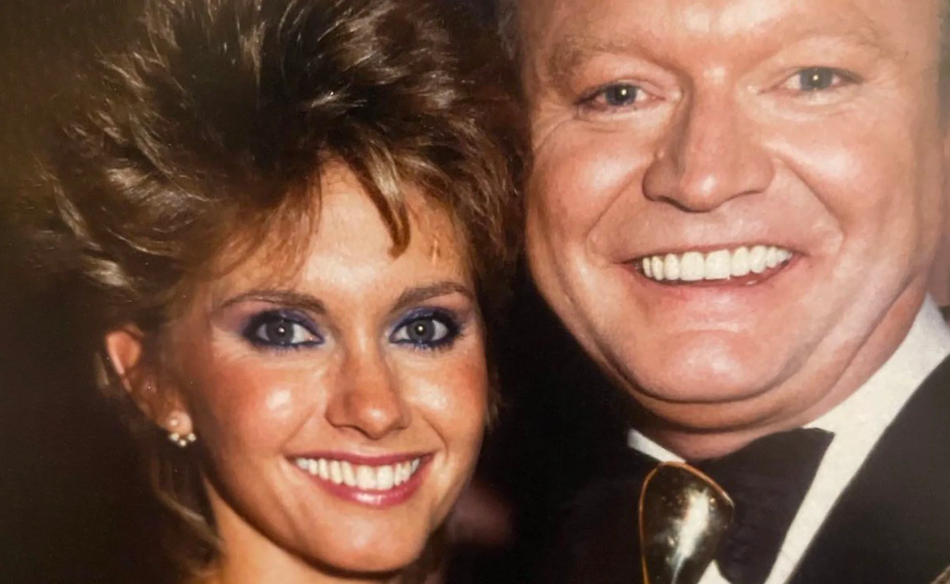 Two smiling individuals pose closely, both wearing formal attire, with one wearing a bow tie.