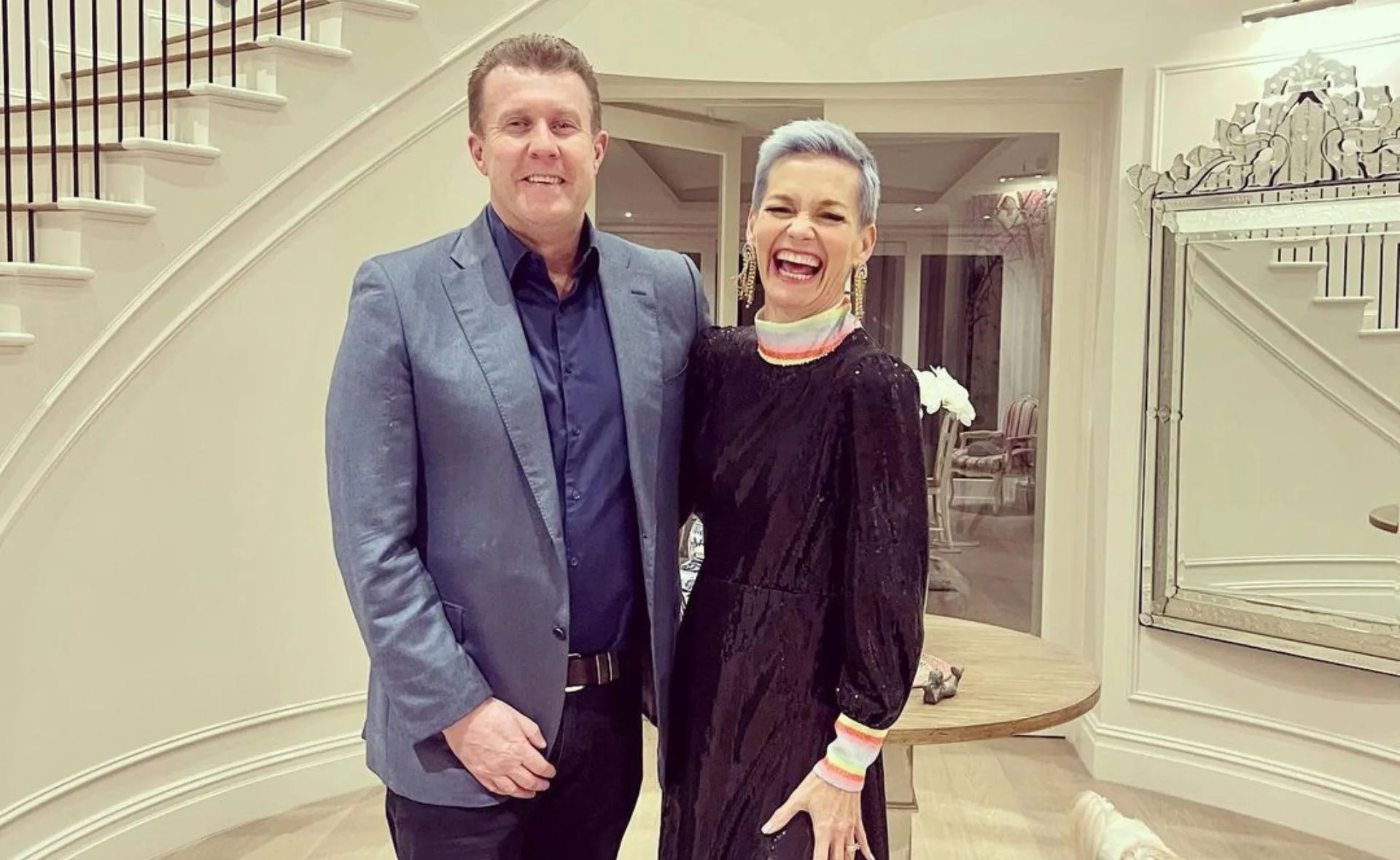 A man in a blue suit and a woman in a black dress with colorful cuffs stand smiling in a stylish hallway.