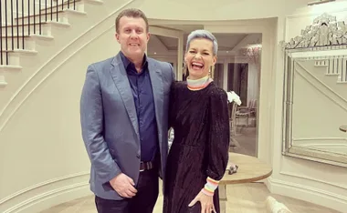 A man in a blue suit and a woman in a black dress with colorful cuffs stand smiling in a stylish hallway.