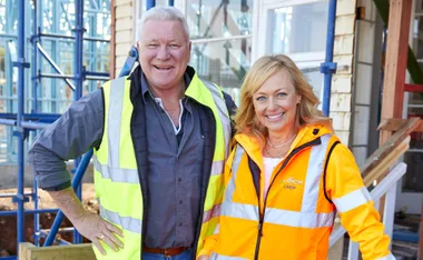 Two people wearing high-visibility jackets smiling on a construction site, scaffolding in the background.