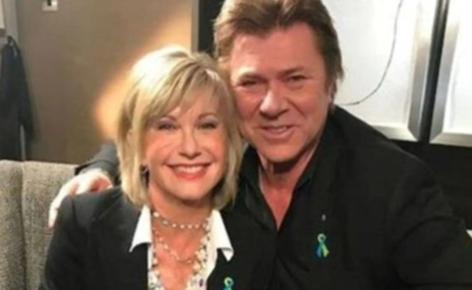 Blonde woman and man in a black suit smiling, seated closely, wearing teal ribbon pins.