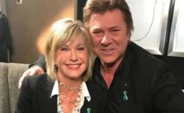 Blonde woman and man in a black suit smiling, seated closely, wearing teal ribbon pins.