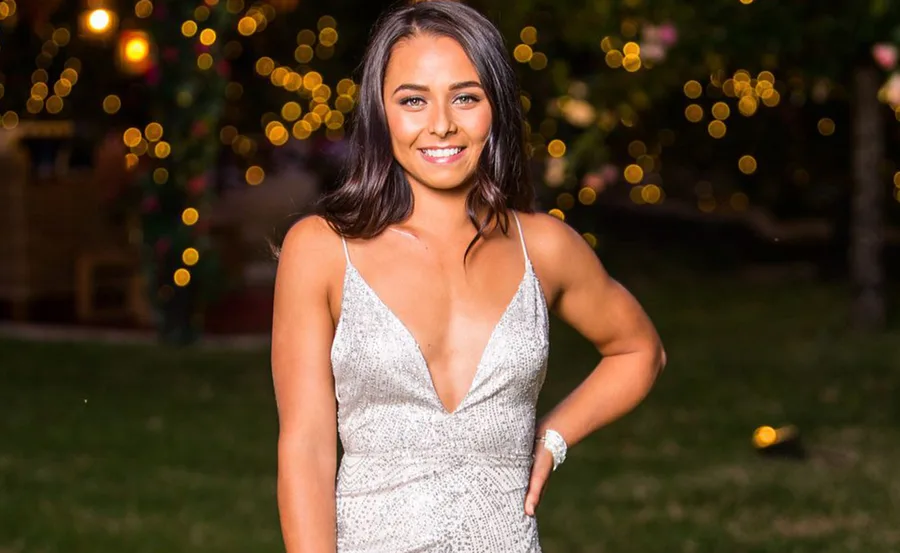 A woman in a sparkly dress stands outside at night, smiling, with blurred lights in the background.