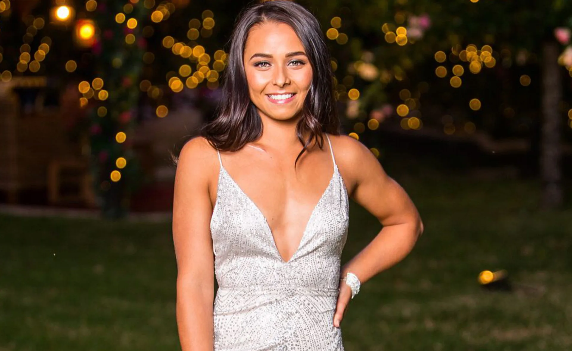 A woman in a sparkly dress stands outside at night, smiling, with blurred lights in the background.
