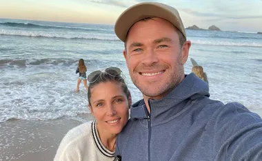 A couple taking a selfie on a beach with waves and children playing in the background.