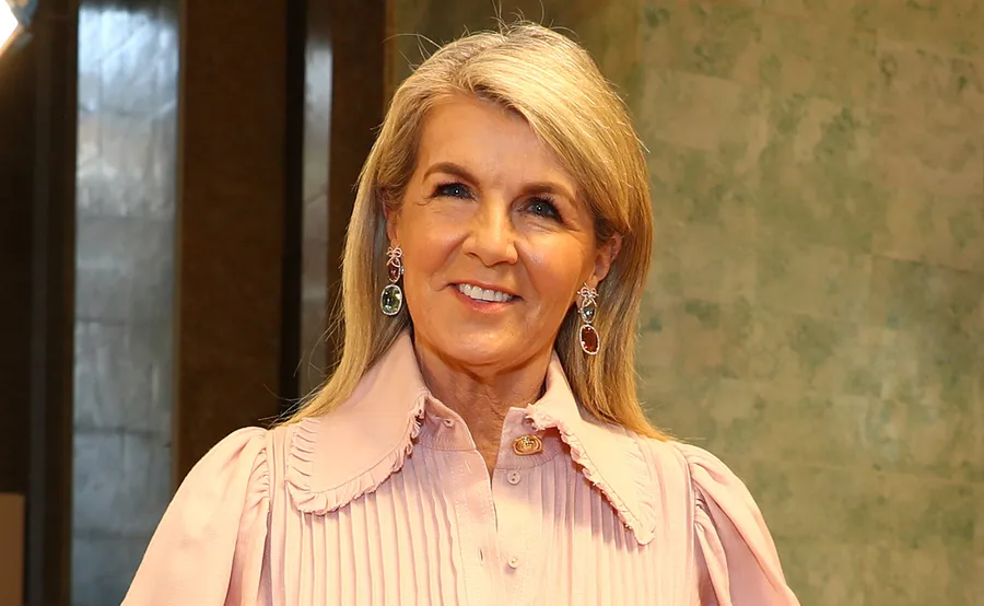 Blonde woman smiling, wearing a pink blouse and colorful earrings, with a textured green wall in the background.