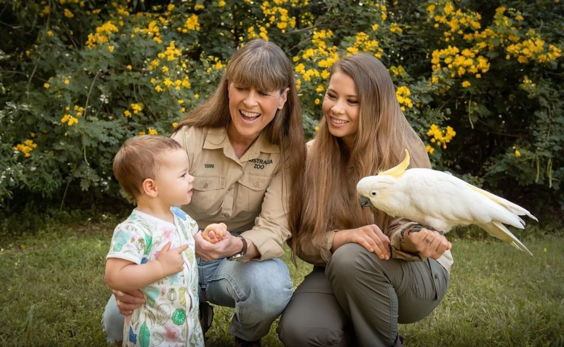 Women and a child outdoors with a white cockatoo perched on one woman's arm, surrounded by yellow flowers.