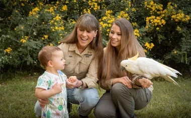 Women and a child outdoors with a white cockatoo perched on one woman's arm, surrounded by yellow flowers.