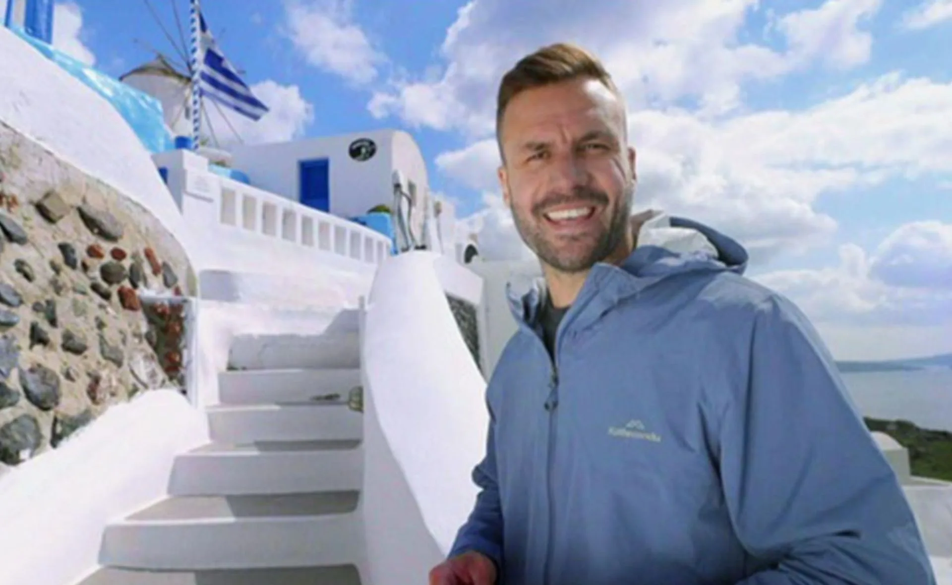 Man smiling in front of white steps and building, with a Greek flag waving in the background under a blue sky.