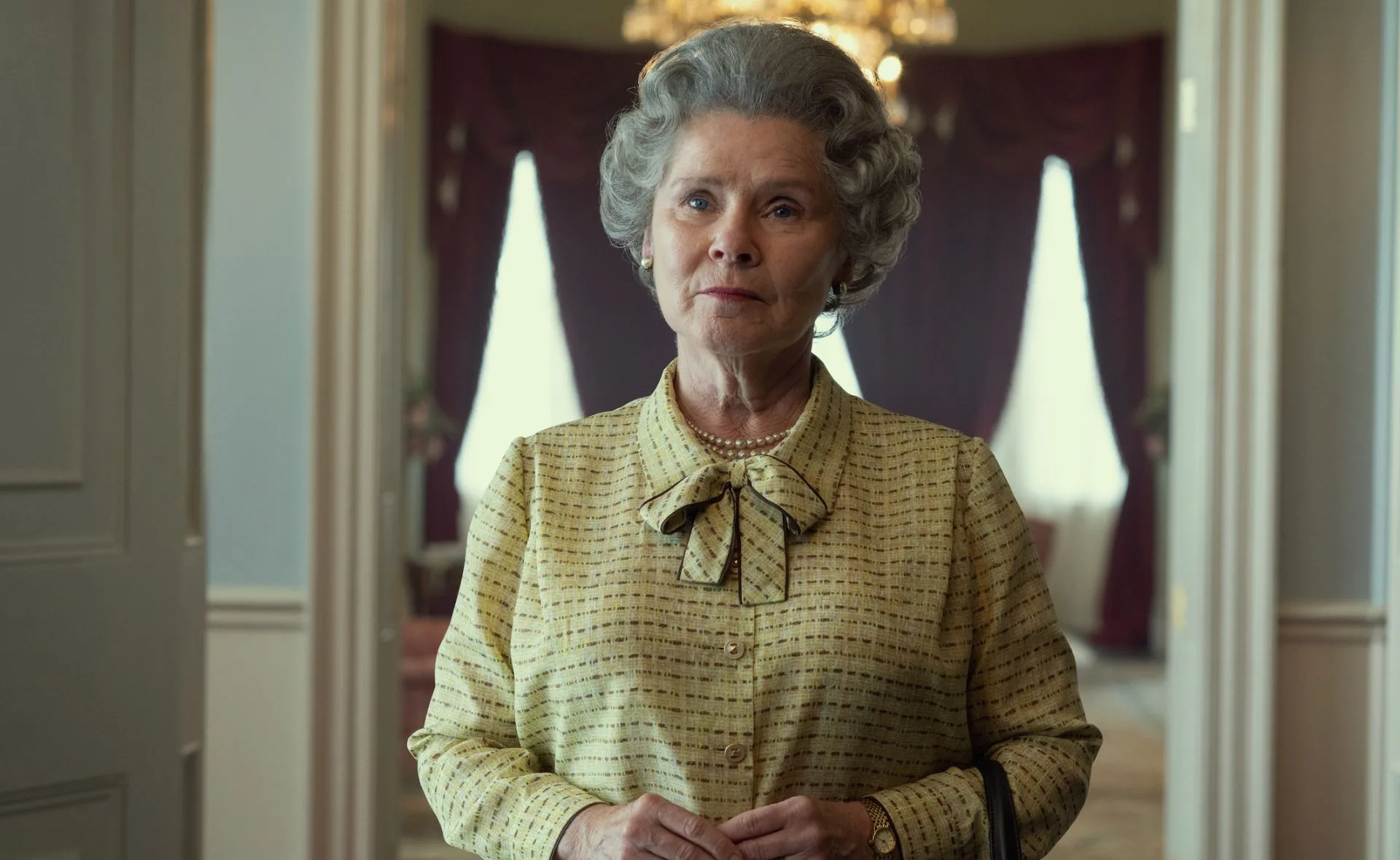 An older woman with grey hair and a bow-tied blouse stands in a well-lit room with curtains and a chandelier in the background.