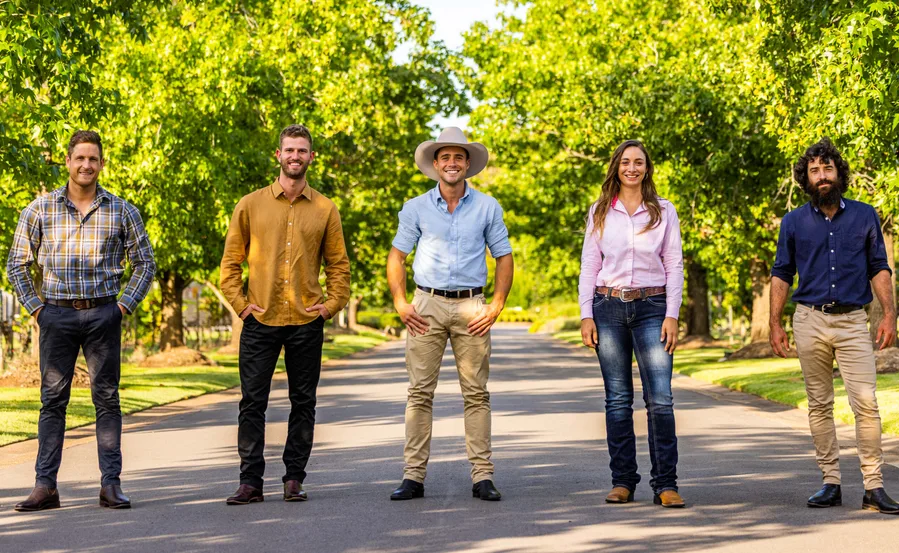 A group of five people stand on a sunny tree-lined road, smiling, dressed in casual and western attire.