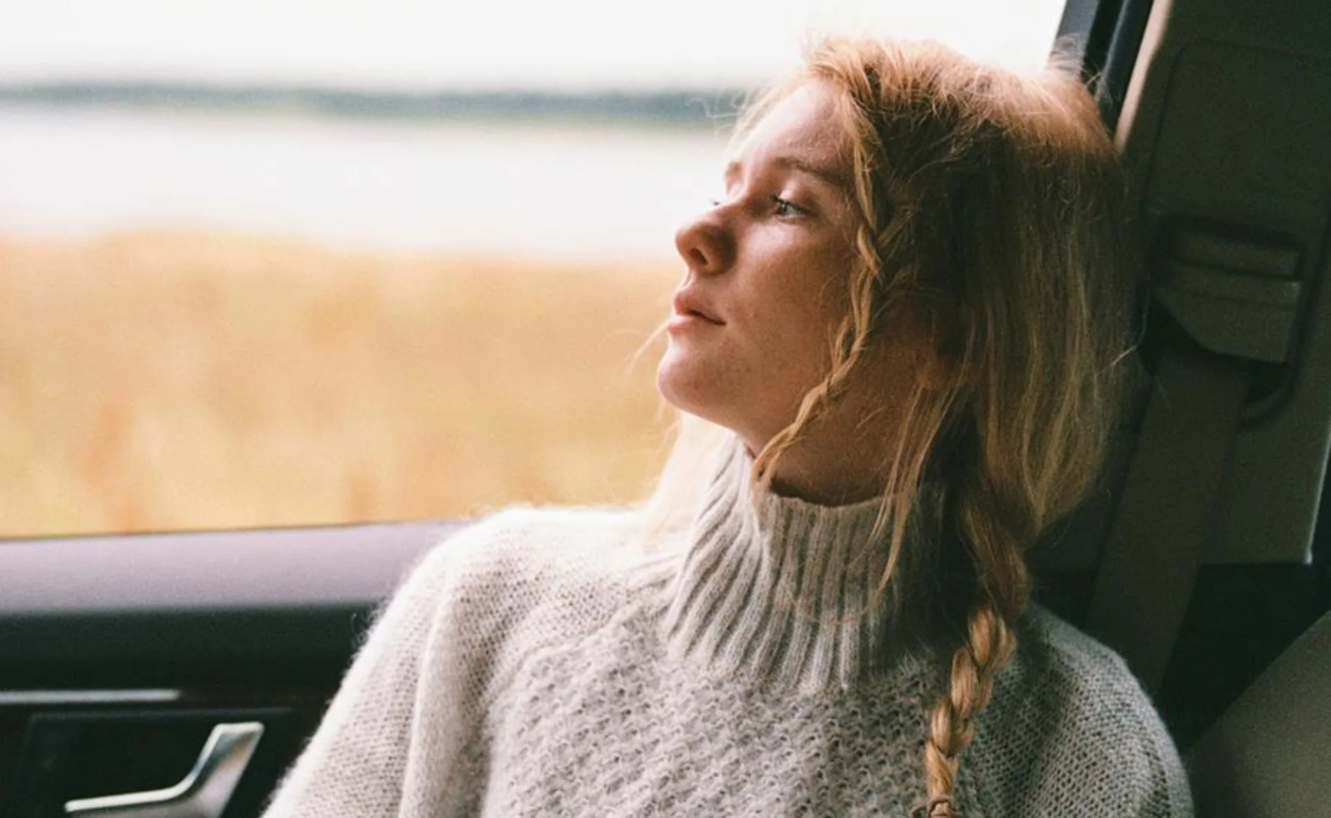 A person with braided hair looks contemplatively out a car window, wearing a cozy sweater, with grassy landscape behind.